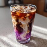 Close-up of a tall glass containing an Iced Lavender Latte with vibrant purple syrup mixing with dark coffee and white cream.