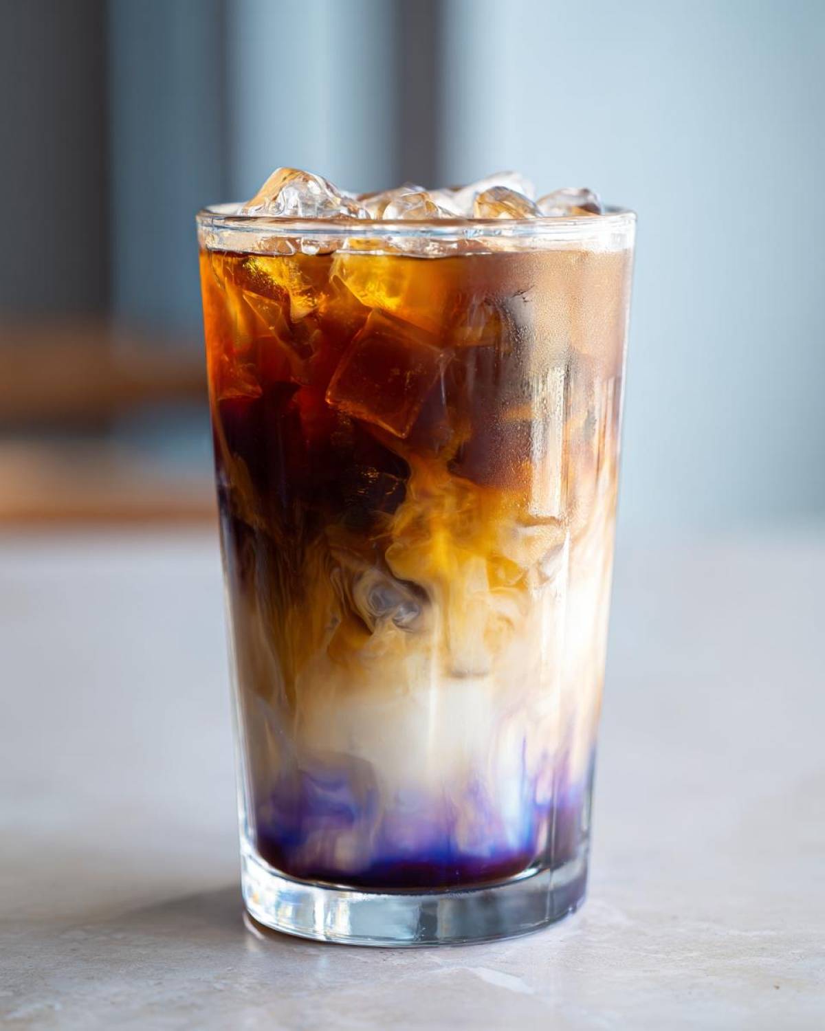 Close-up of a tall glass filled with an Iced Lavender Latte, showing swirling layers of dark coffee, white milk, and purple lavender syrup over ice.