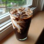Close-up of a tall glass containing a rich Iced Mocha with milk swirling dramatically into the dark coffee and chocolate base, topped with ice.