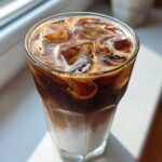 Close-up of a tall glass containing a layered Iced Mocha with ice cubes and swirling cream.