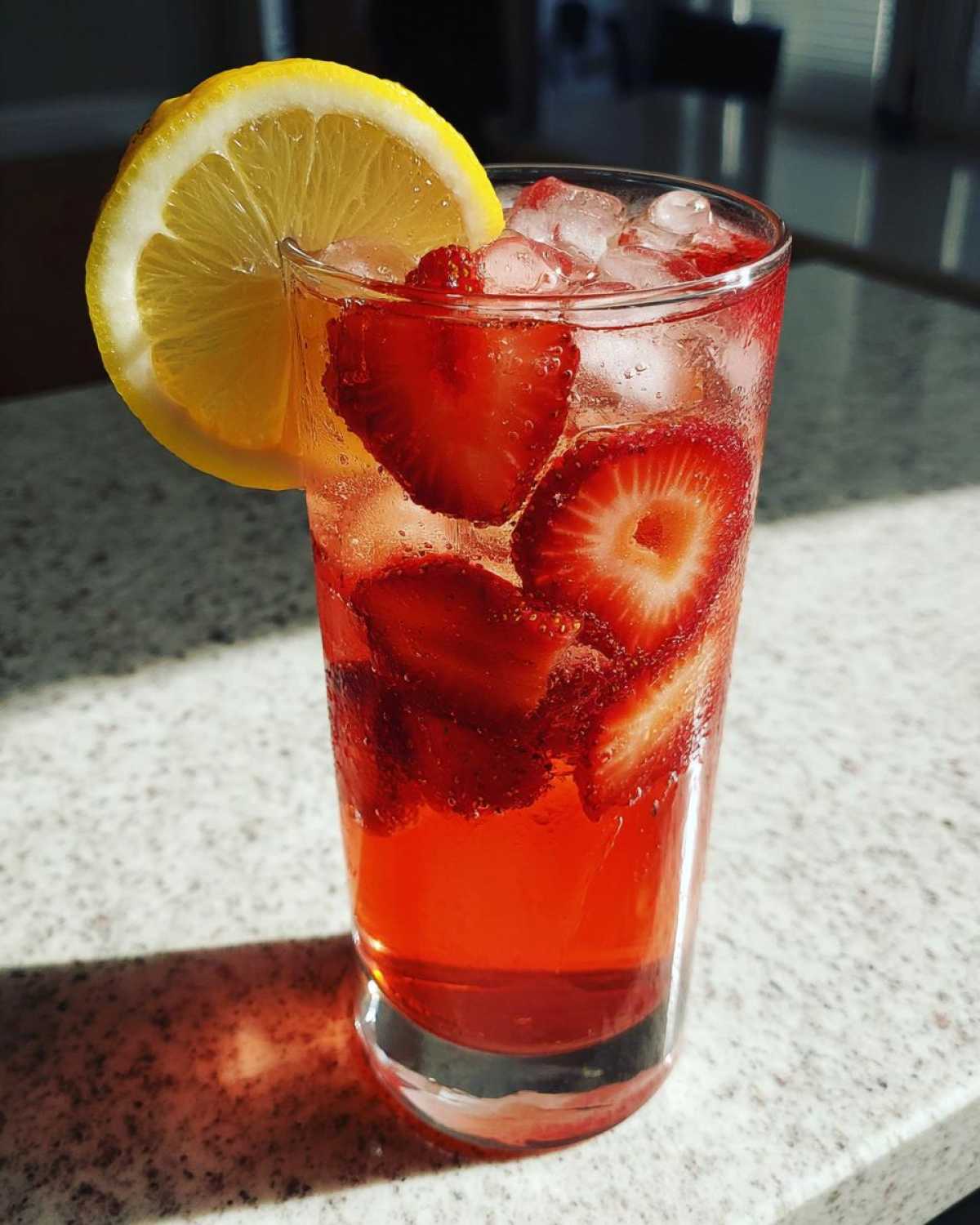 A tall glass filled with Iced Strawberry Lemon Tea, ice, sliced strawberries, and a lemon wedge garnish.