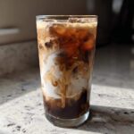 A tall glass filled with an Iced Vanilla Chai Latte showing rich swirls of cream mixing with dark chai concentrate and ice.