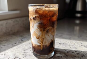 A tall glass filled with an Iced Vanilla Chai Latte showing rich swirls of cream mixing with dark chai concentrate and ice.