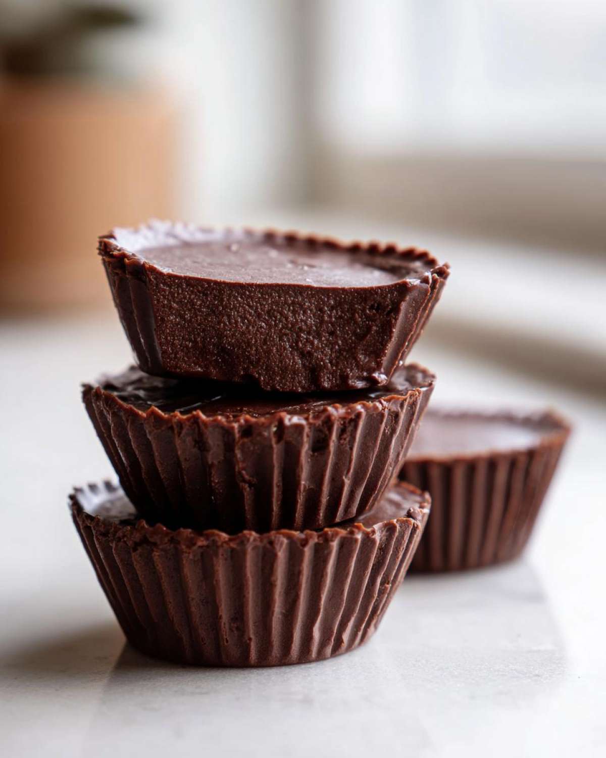 A stack of three rich, dark chocolate Keto Chocolate Peanut Butter Fat Bombs in fluted paper cups.