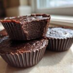 A close-up stack of rich, dark chocolate Keto Chocolate Peanut Butter Fat Bombs in brown paper liners.