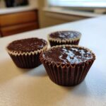Three freshly made Keto Chocolate Peanut Butter Fat Bombs sitting in brown paper liners on a white countertop.