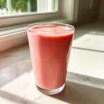 A tall glass filled with a thick, pink Keto Strawberry Cheesecake Smoothie sitting on a white marble surface near a window.