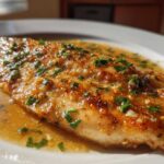 A close-up of a perfectly cooked Lemon Garlic Butter Tilapia fillet swimming in savory sauce and topped with fresh parsley.