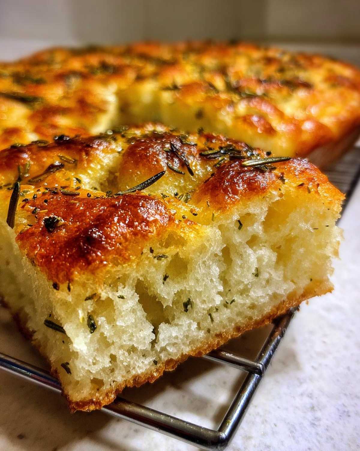 A close-up view of a thick slice of No-Knead Skillet Focaccia with Rosemary showing its airy interior and golden, herb-topped crust.