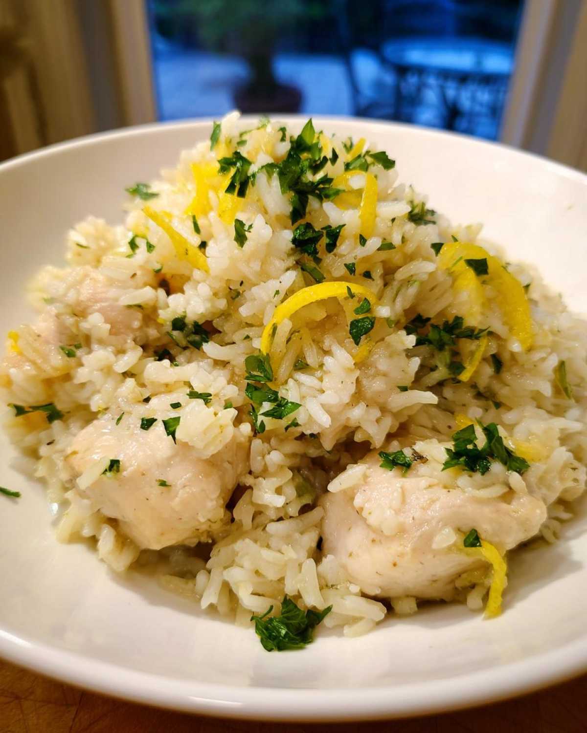 A close-up of One-Pot Chicken and Rice with Lemon and Herbs garnished with fresh parsley and lemon zest in a white bowl.