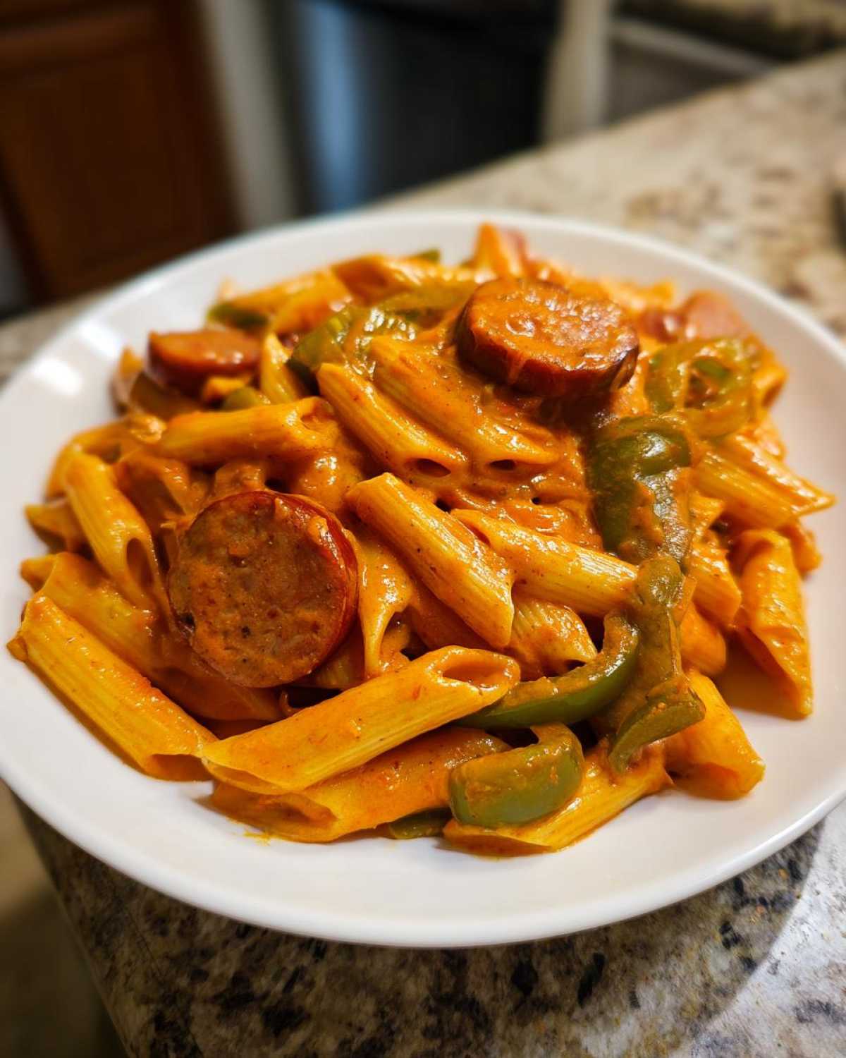 A close-up of a white bowl filled with One-Pot Creamy Cajun Sausage Pasta, featuring penne, sliced sausage, and green peppers.