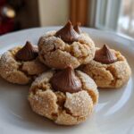 A close-up of four delicious Peanut Butter Blossom Cookies, rolled in sugar and topped with a chocolate kiss, resting on a white plate.