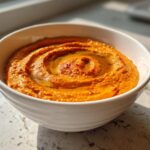 A close-up of bright orange Roasted Red Pepper Hummus swirled in a white bowl, drizzled with olive oil.