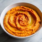 Close-up of bright orange Roasted Red Pepper Hummus swirled in a white bowl, drizzled with olive oil and sprinkled with paprika.
