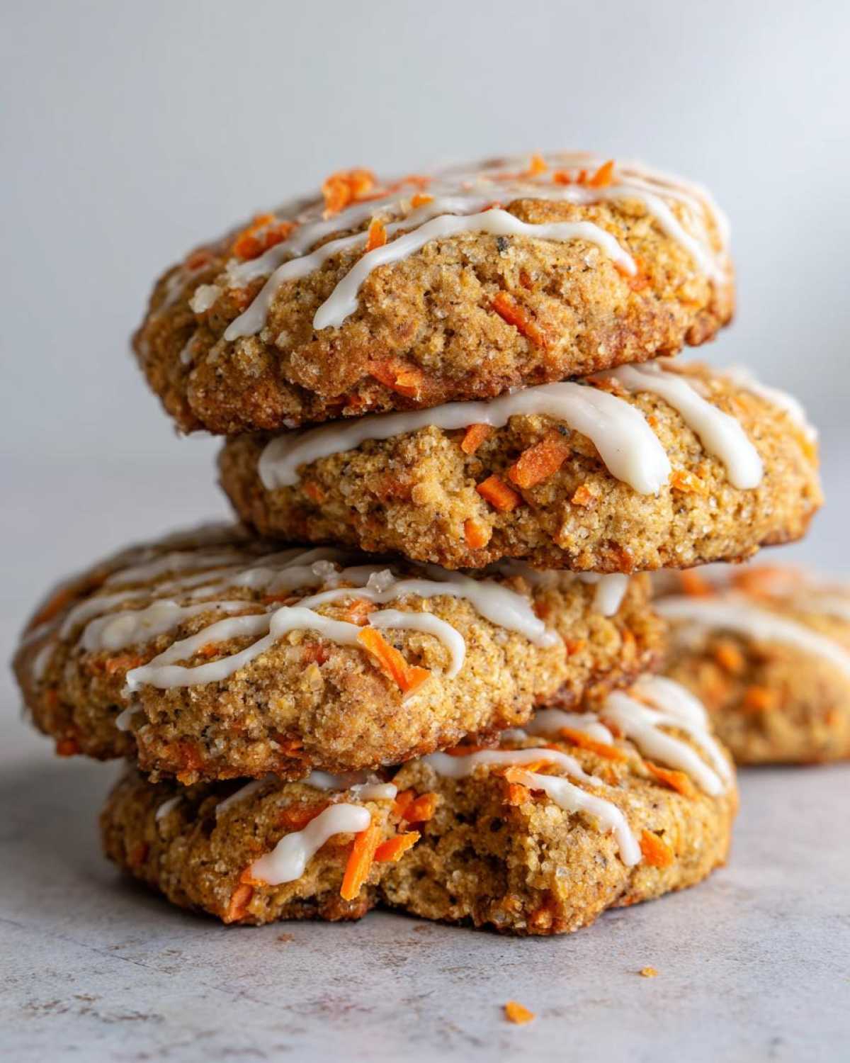 A stack of four Soft-Baked Carrot Cake Cookies generously drizzled with cream cheese glaze and visible shredded carrots.