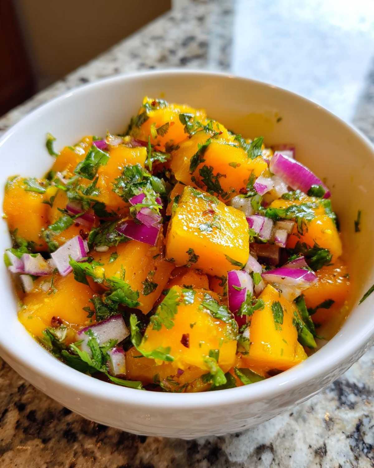 Close-up of bright orange chunks of mango mixed with diced red onion and fresh cilantro in a white bowl, showcasing the Spicy Mango Salsa.