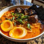 A vibrant bowl of Spicy Miso Ramen featuring rich orange broth, soft-boiled eggs cut in half, beef chunks, and nori.