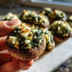A hand holding one perfectly baked Spinach and Feta Stuffed Mushroom, showing the cheesy filling.