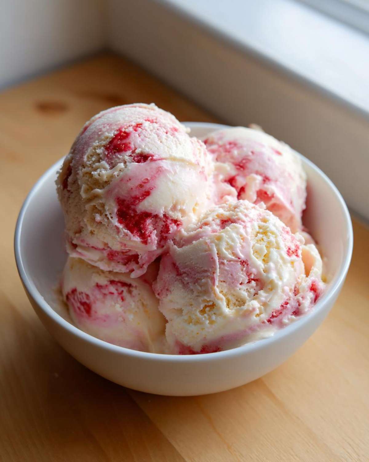Four scoops of creamy Strawberry Banana Ice Cream swirled with bright pink strawberry puree in a white bowl.