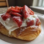Close-up of a flaky Strawberry Cream Cheese Danish topped with whipped cream and sliced strawberries.