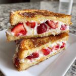 Two halves of golden-brown Strawberry Cream Cheese Stuffed French Toast stacked on a white plate.