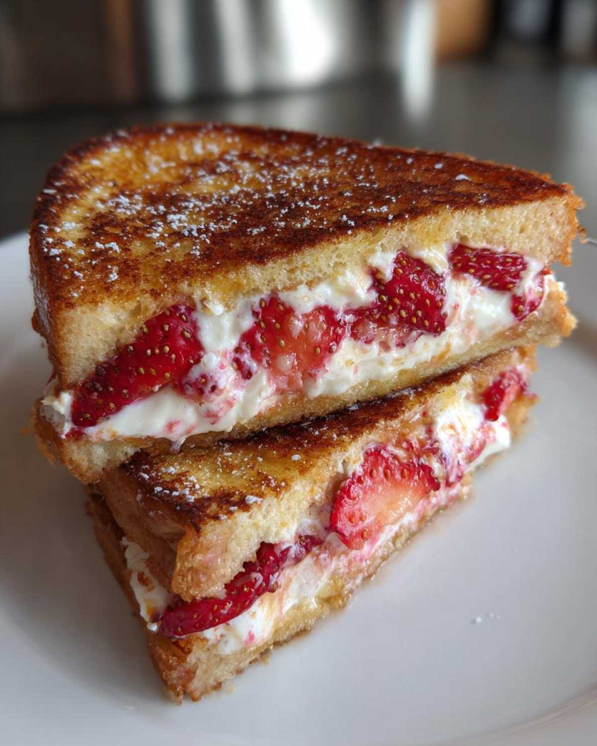 Two halves of golden brown Strawberry Cream Cheese Stuffed French Toast stacked on a white plate, dusted with powdered sugar.