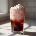 A tall glass of Strawberry Cream Cold Brew with iced coffee base, pink foam, and whipped cream topping.