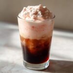 A tall glass filled with layered Strawberry Cream Cold Brew, featuring dark coffee below and thick pink foam topping.