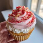 A hand holding a delicious Strawberry Crunch Cupcakes topped with pink frosting and crunchy red and tan sprinkles.