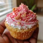 A close-up of a perfectly frosted Strawberry Crunch Cupcakes, topped with pink frosting and crunchy red sprinkles, held by a hand.