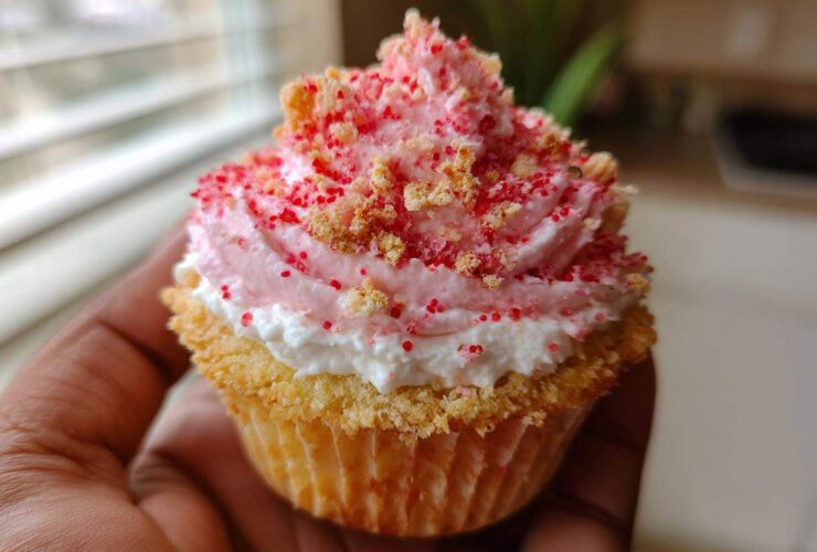 A close-up of a perfectly frosted Strawberry Crunch Cupcakes, topped with pink frosting and crunchy red sprinkles, held by a hand.