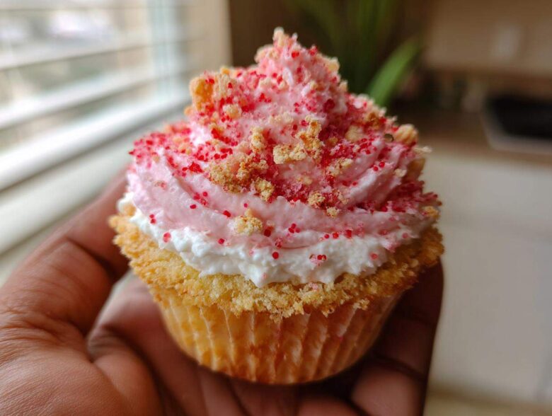 A close-up of a perfectly frosted Strawberry Crunch Cupcakes, topped with pink frosting and crunchy red sprinkles, held by a hand.