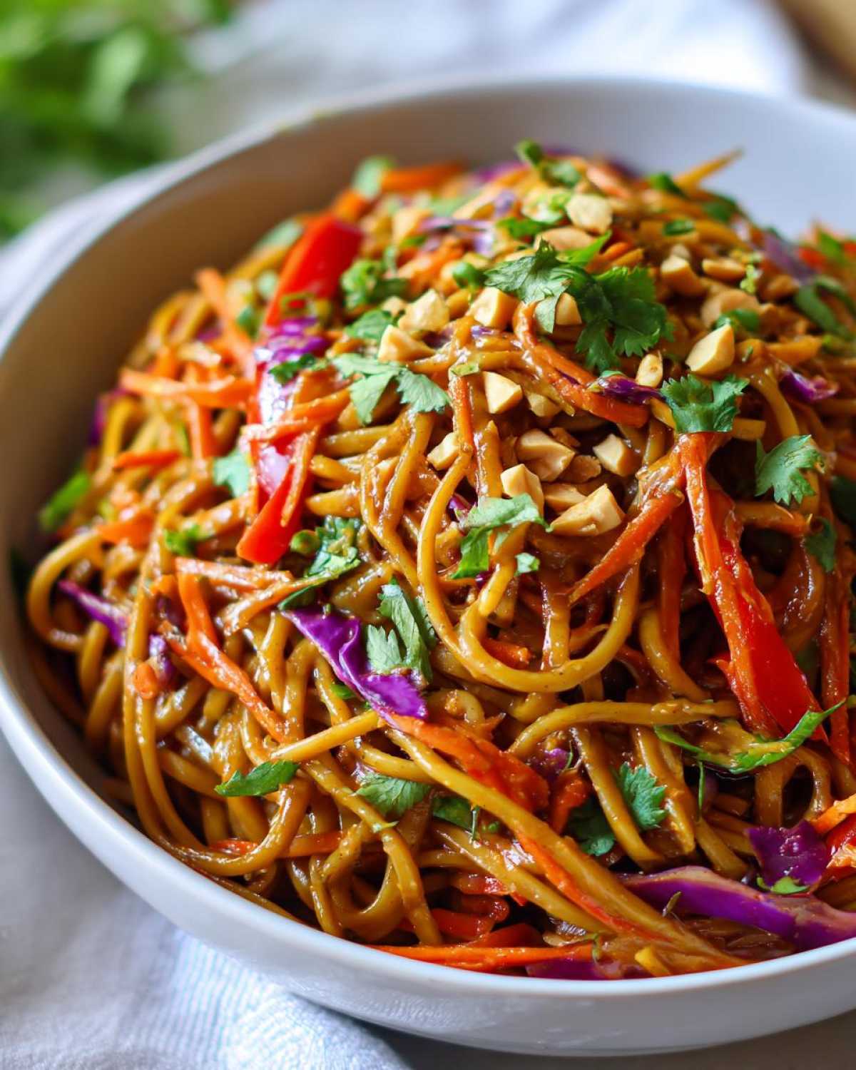 Close-up of a bowl filled with saucy Thai Peanut Noodles with Crunchy Veggies, topped with peanuts and cilantro.