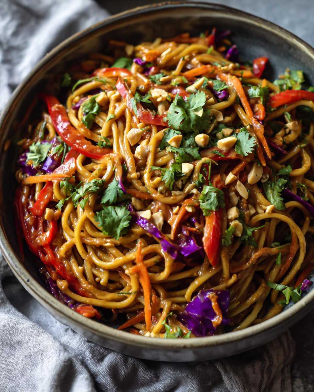 A close-up bowl of vibrant Thai Peanut Noodles with crunchy veggies like red peppers, carrots, and purple cabbage, topped with cilantro and peanuts.