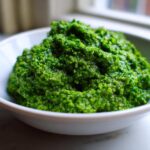 A close-up of vibrant green, textured Vegan Basil Pesto piled high in a small white bowl.