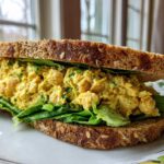 A close-up of a hearty Vegan Chickpea Salad Sandwich layered with bright yellow filling and crisp lettuce on seeded brown bread.