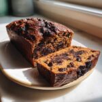 A loaf of rich Vegan Chocolate Banana Bread with one thick slice cut and resting beside it on a light plate.