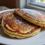 A stack of three fluffy Vegan Lemon Poppy Seed Pancakes dusted with powdered sugar on a white plate.