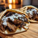 Two servings of Vegan Mushroom Shawarma stuffed into warm pita bread and drizzled with white sauce, resting on a wooden cutting board.
