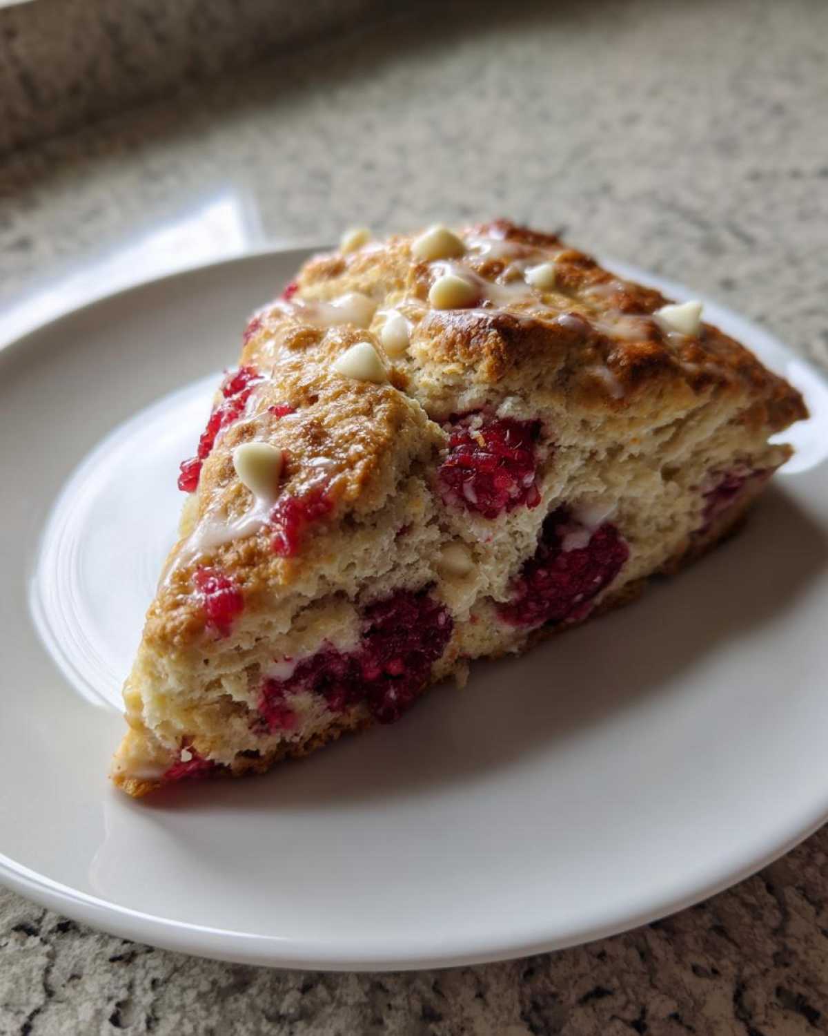 A triangular slice of White Chocolate Raspberry Scones topped with white chocolate chips and a light glaze, served on a white plate.