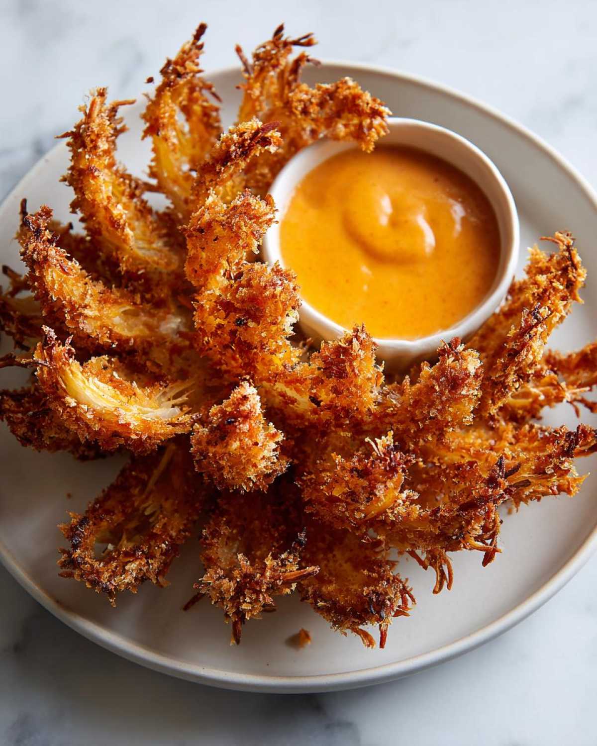 A whole, deep-fried Air Fryer Crispy Onion Petals served on a plate with a small bowl of orange spicy mayo dipping sauce.