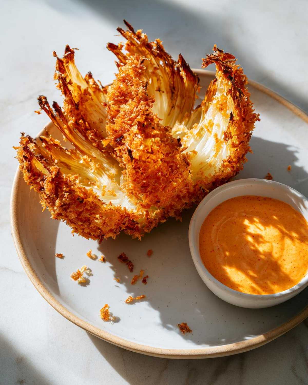 A whole, blooming Air Fryer Crispy Onion Petals served on a plate next to a small bowl of orange spicy mayo dip.