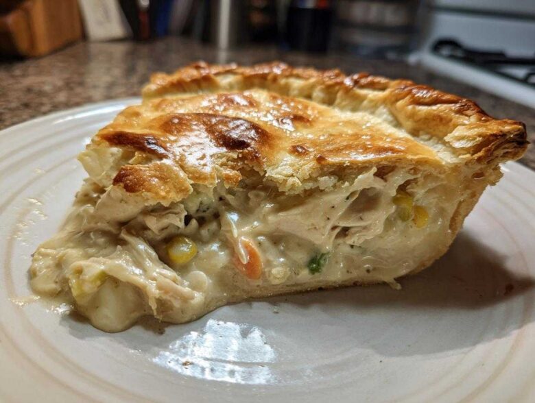 A close-up of a generous slice of chicken pot pie recipe showing creamy filling, shredded chicken, corn, and carrots under a golden, flaky crust.