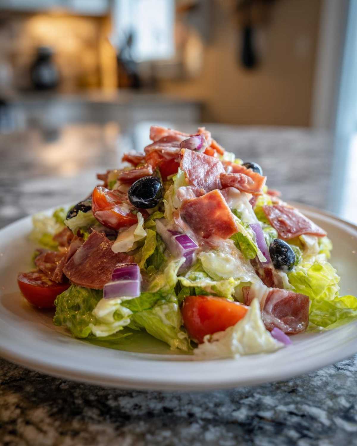 A vibrant, towering serving of grinder salad featuring chopped lettuce, salami, tomatoes, red onion, and black olives, drizzled with dressing.