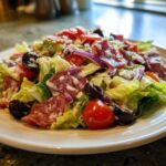 A vibrant, creamy grinder salad featuring crisp lettuce, salami slices, black olives, red onion, and tomatoes on a white plate.