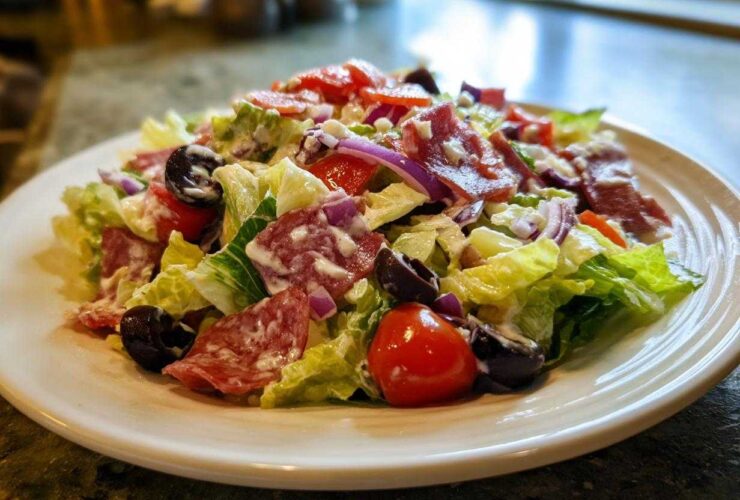A vibrant, creamy grinder salad featuring crisp lettuce, salami slices, black olives, red onion, and tomatoes on a white plate.