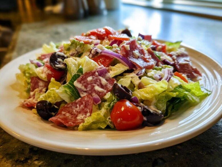 A vibrant, creamy grinder salad featuring crisp lettuce, salami slices, black olives, red onion, and tomatoes on a white plate.
