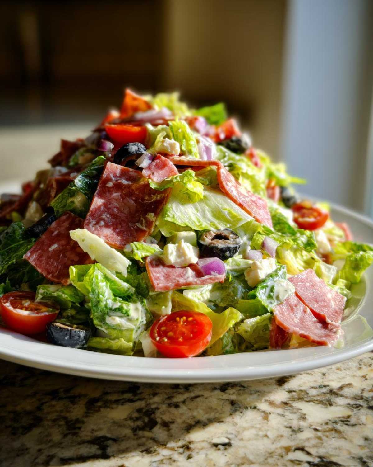 A tall mound of vibrant grinder salad featuring lettuce, salami, cherry tomatoes, olives, and cheese, tossed in dressing.
