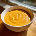 A close-up of a thick, vibrant orange cheese sauce swirled perfectly in a white bowl.