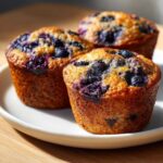 Close-up of three golden-brown blueberry cottage cheese muffins bursting with dark blueberries on a white plate.